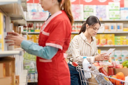 Photo for Young female customer leaning on a full shopping cart and checking a shopping list at the supermarket, a stock clerk is working in the foreground - Royalty Free Image