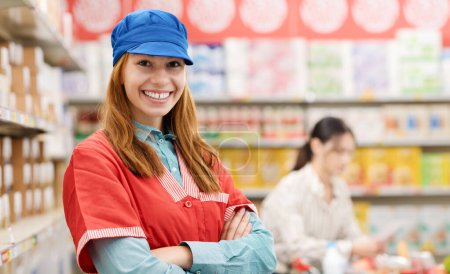 Téléchargez les photos : Jeune vendeuse confiante posant les bras croisés au supermarché, elle sourit à la caméra - en image libre de droit