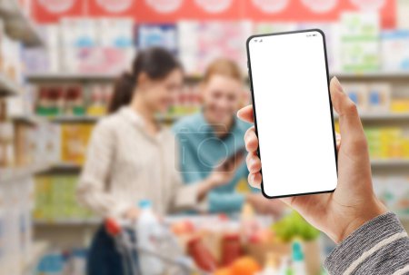 Téléchargez les photos : Femme tenant un smartphone avec écran vierge et intérieur de supermarché en arrière-plan, concept d'épicerie en ligne - en image libre de droit
