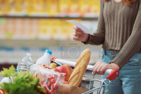 Photo for Young woman doing grocery shopping at the supermarket, she is pushing a full trolley and holding a shopping list, close up - Royalty Free Image