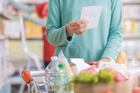 Téléchargez les photos : Client achetant des produits d'épicerie au supermarché, elle tient une liste d'épicerie et pousse un panier complet - en image libre de droit