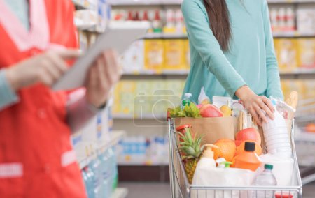 Téléchargez les photos : Femme poussant un panier complet au supermarché et commis de stock au travail, concept intérieur de supermarché - en image libre de droit
