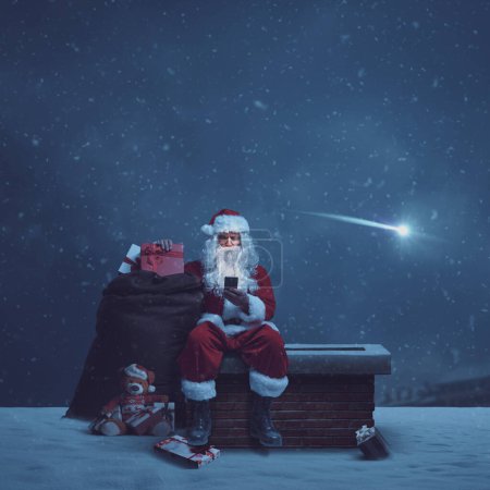 Foto de Santa Claus entrega regalos en Nochebuena, él está sentado en una chimenea y conectarse en línea con su teléfono inteligente - Imagen libre de derechos