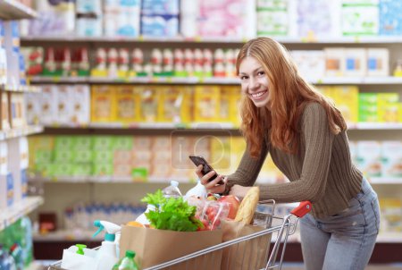 Téléchargez les photos : Portrait d'une jeune femme faisant de l'épicerie au supermarché, elle sourit et tient son smartphone - en image libre de droit