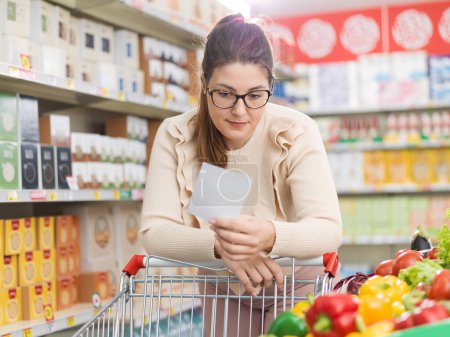 Foto de Mujer haciendo compras de comestibles en el supermercado en la sección de productos, ella se apoya en el carro de la compra y sostiene una lista de supermercados - Imagen libre de derechos