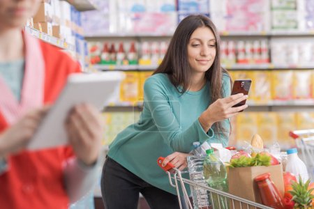 Foto de Mujer joven usando un teléfono inteligente en el supermercado y sonriendo a la cámara - Imagen libre de derechos