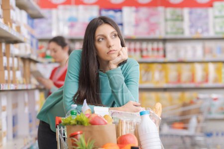 Photo for Bored young woman leaning on the shopping cart at the grocery store, she is looking around and feeling tired - Royalty Free Image