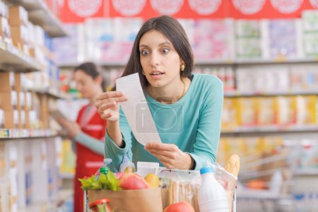 Foto de Joven mujer sorprendida en el supermercado, ella se apoya en un carro de la compra y comprobar un recibo de comestibles caro largo - Imagen libre de derechos