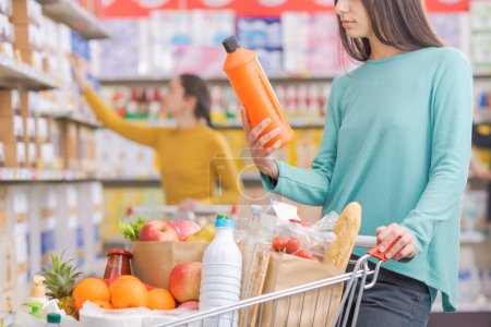 Photo for Woman buying products at the grocery store, she is holding a detergent bottle and leaning on a full shopping cart - Royalty Free Image