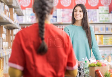 Foto de Mujer sonriente comprando comestibles en el supermercado, ella está empujando un carrito de compras y hablando con un asistente de tienda amigable, concepto de compras de comestibles - Imagen libre de derechos