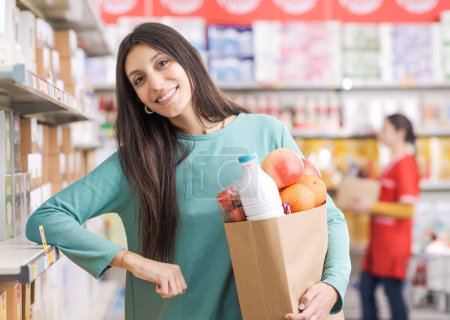 Téléchargez les photos : Portrait d'une jeune femme heureuse tenant un sac plein d'épicerie au supermarché, elle sourit à la caméra - en image libre de droit