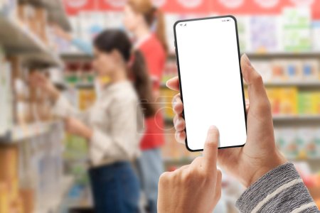Foto de Mujer sosteniendo un teléfono inteligente con pantalla en blanco y el interior del supermercado en el fondo, concepto de compra de comestibles en línea - Imagen libre de derechos