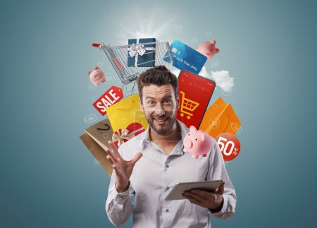 Téléchargez les photos : Homme heureux excité tenant une tablette et faisant des achats en ligne, il est entouré d'articles d'achat, d'offres et de concept de vente - en image libre de droit