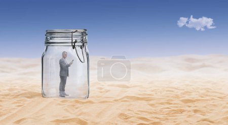 Foto de Empresario de pie dentro de un gran frasco y utilizando un teléfono inteligente, está atrapado y perdido en el desierto - Imagen libre de derechos