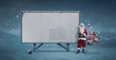 Foto de Compras de Navidad: cartel publicitario en blanco y Santa junto a un carrito de compras completo - Imagen libre de derechos