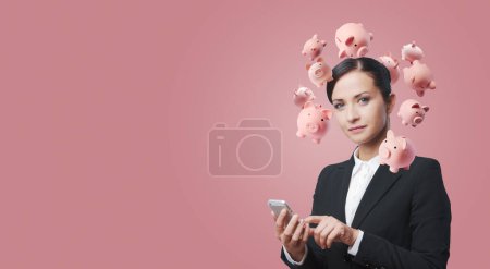 Téléchargez les photos : Femme d'affaires confiante utilisant un smartphone et planifiant ses investissements, elle a de nombreuses tirelires autour de la tête - en image libre de droit