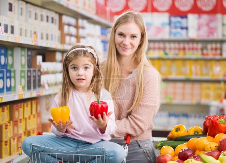 Téléchargez les photos : Jolie fille mignonne assise sur un panier et achetant des légumes et des fruits frais avec sa mère au supermarché - en image libre de droit