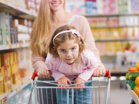 Photo for Happy girl doing grocery shopping with her mom, she is leaning on the shopping cart and smiling - Royalty Free Image