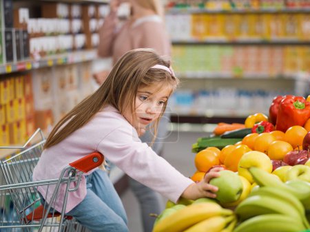 Foto de Linda chica encantadora sentada en el carrito de la compra, ella está tomando una fruta en la sección de productos mientras su madre está ocupada tomando productos en los estantes en el fondo - Imagen libre de derechos