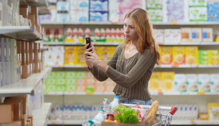 Téléchargez les photos : Jeune femme faisant des courses au supermarché, elle vérifie les informations alimentaires à l'aide d'une application de scanner de nourriture sur son smartphone - en image libre de droit