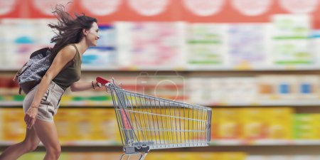 Photo for Happy young woman doing grocery shopping, she is running and pushing a shopping cart, copy space - Royalty Free Image
