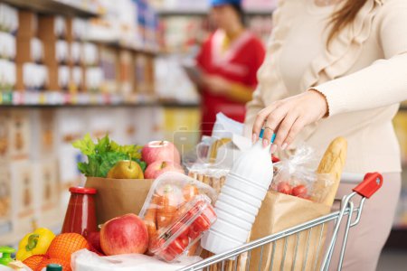 Téléchargez les photos : Femme achetant des produits frais au supermarché, elle pousse un panier plein et tient une bouteille de lait - en image libre de droit
