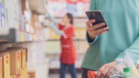 Téléchargez les photos : Femme utilisant un smartphone au supermarché et faisant des courses, mains fermées - en image libre de droit