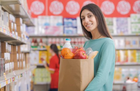 Foto de Mujer joven sonriente sosteniendo una bolsa llena de comestibles y mirando una cámara, empleado de stock trabajando y estantes de supermercados en el fondo - Imagen libre de derechos