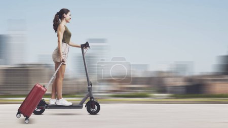 Photo for Young woman riding an electric scooter and travelling through the city, she is carrying a trolley bag - Royalty Free Image