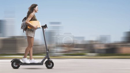 Foto de Estudiante sosteniendo cuadernos y montando un scooter eléctrico, paisaje urbano en el fondo - Imagen libre de derechos
