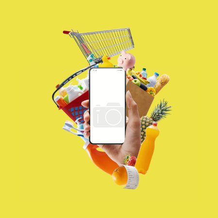 Foto de Mano sosteniendo un teléfono inteligente con pantalla en blanco rodeado de comestibles y artículos de compras, concepto de compras en línea - Imagen libre de derechos