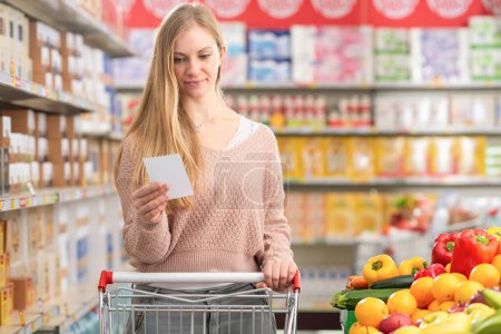 Téléchargez les photos : Jolie jeune femme poussant un panier et achetant des produits d'épicerie au supermarché - en image libre de droit