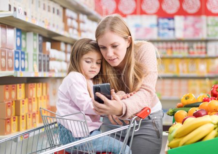 Téléchargez les photos : Jolie fille assise sur un panier d'achat au supermarché, sa mère montre ses applications sur le smartphone : concept de technologie et d'épicerie - en image libre de droit