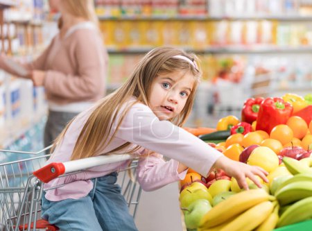 Téléchargez les photos : Jolie fille surprise prenant un fruit dans la section des produits au supermarché, elle est assise sur le panier, concept de nourriture saine - en image libre de droit