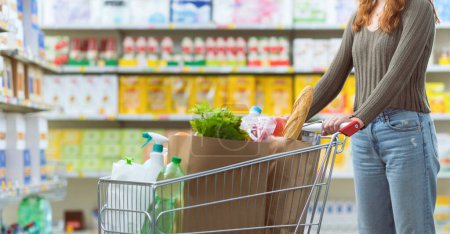 Téléchargez les photos : Femme cliente achetant des produits d'épicerie au supermarché, elle pousse un panier complet - en image libre de droit