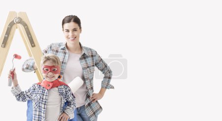 Téléchargez les photos : Joyeux jeune mère et garçon souriant peignant leur maison, l'enfant porte un costume de super héros - en image libre de droit