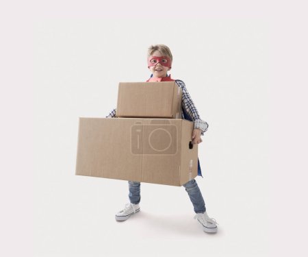 Foto de Lindo chico superhéroe con máscara y capa, que está llevando fácilmente cajas pesadas gracias a sus superpoderes - Imagen libre de derechos