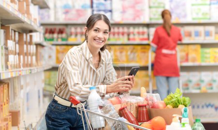 Téléchargez les photos : Femme souriante poussant un panier complet et utilisant un smartphone au supermarché, épicerie et concept de technologie - en image libre de droit