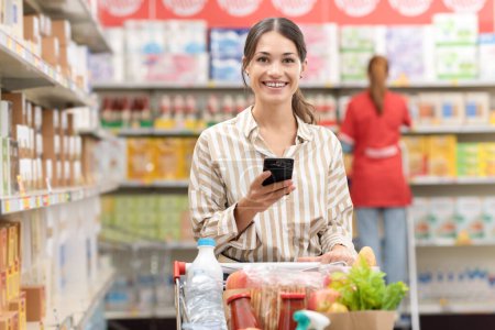 Foto de Mujer sonriente empujando un carrito de compras completo y utilizando un teléfono inteligente en el supermercado, compras de comestibles y el concepto de tecnología - Imagen libre de derechos