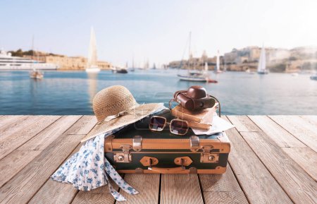Téléchargez les photos : Valise de style vintage et accessoires sur une terrasse en bois, port de ville en arrière-plan : concept de destinations de voyage et vacances - en image libre de droit