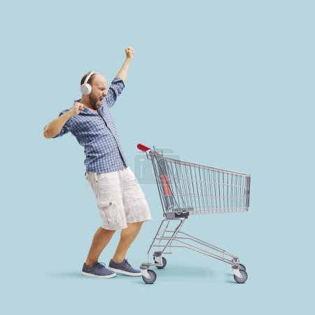 Foto de Emocionado hombre haciendo compras de comestibles, está escuchando música y bailando al lado de un carrito de compras vacío - Imagen libre de derechos