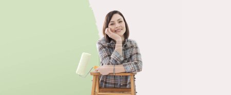 Téléchargez les photos : Heureuse jeune femme peignant des murs dans sa nouvelle maison, elle tient un rouleau de peinture et sourit, espace de copie - en image libre de droit