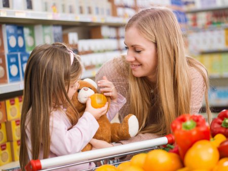 Téléchargez les photos : Mère et fille faisant des courses ensemble, la fille tient un fruit et nourrit sa peluche - en image libre de droit