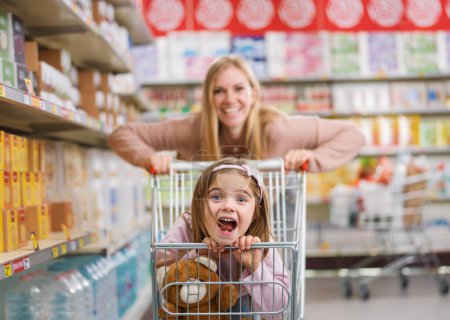 Foto de Chica linda alegre sentada en un carrito de compras en el supermercado, su madre la está empujando: compras de comestibles y concepto de estilo de vida - Imagen libre de derechos