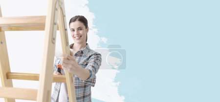 Foto de Joven mujer sonriente pintando su casa, ella está de pie junto a una escalera y sosteniendo un rodillo de pintura - Imagen libre de derechos