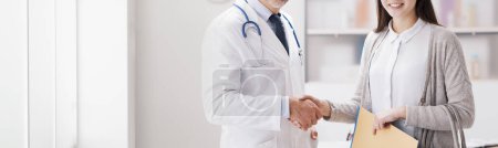 Téléchargez les photos : Souriant médecin confiant serrant la main d'une patiente dans le bureau, elle détient des dossiers médicaux - en image libre de droit