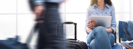 Téléchargez les photos : Jeune voyageuse assise dans la salle d'attente du terminal, elle se connecte à une tablette à écran tactile et au réseautage - en image libre de droit