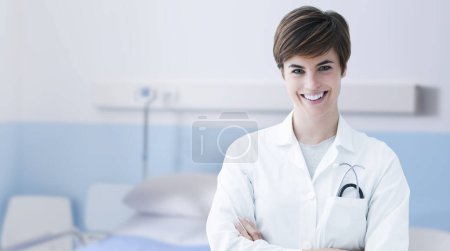 Foto de Joven doctora sonriente posando con los brazos cruzados en una habitación de hospital, asistencia médica y concepto de atención médica - Imagen libre de derechos