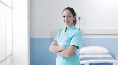 Foto de Enfermera joven y confiada que trabaja en el hospital, posa con los brazos cruzados y sonriendo - Imagen libre de derechos