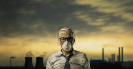 Photo for Hopeless dirty businessman wearing a face mask and looking at camera, polluting industrial plant and toxic clouds in the background - Royalty Free Image